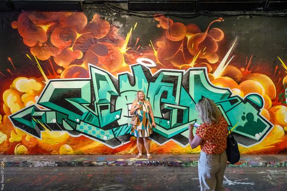 A woman posing in front of a green graffiti tag surrounded by orange flames while her friend reviews the photo she's just taken.