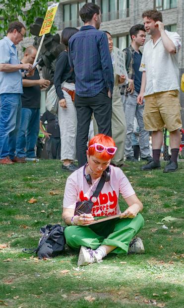 A young woman with red hair and sun glasses on her head sits on the grass amidst the protest and writes a slogan on her sign