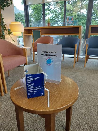 Photo of a waiting area in a Dr's office. A box of masks sits on a table with a sign who's text reads: if you think you are sick or have a cold, then please wear a mask.