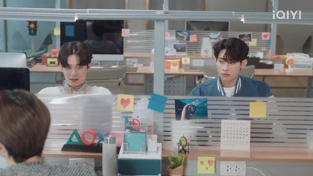 Jade and Mai are seated at their workstations in the office. Jade is wearing glasses, a scarf and a white sweater, while Mai is in a blue and white varsity jacket. Their desks are decorated with colorful sticky notes and office supplies.

Mai is looking to the side at Jade, with a wounded pout, while Jade looks down at his desk, unaware.