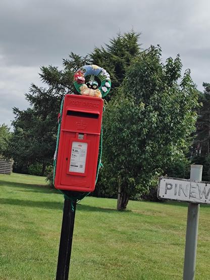 A photo of a crochet summer wreath on top of a red post box on a pole next to a grass verge. The wreath is green, yellow and blue and has crochet flowers, sheep, dogs , a tractor and a sun on it.