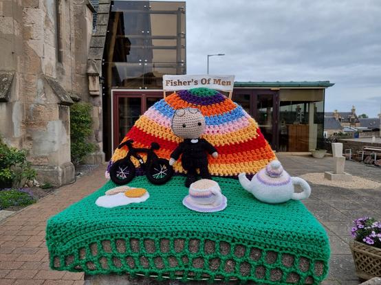 A photo of a concrete plinth with a crochet scene on it in front of a church. The surface is green and it has a rainbow hill behind it with the sign "Fishers of Men" in gold lettering on a white sign. Underneath this is a crochet vicar dressed in black with a white dog collar and little round glasses on his head. He is standing next to a crochet bicycle and there is a teapot and cup and some crochet biscuits in front of him.