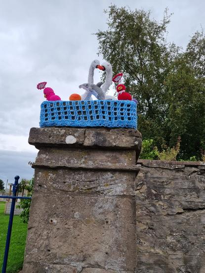 A photo of a concrete plinth with a blue basket crocheted on the top with two crochet swans, a dolphin , a pink and red bird and a ball