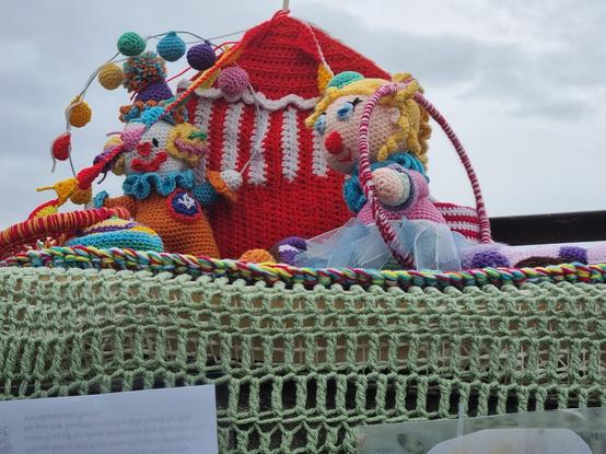 A photo of a post that has been yarn bombed with a crochet scene of a big top tent, behind which is a multicoloured big wheel and in front of which are two colourful crochet clowns. The one nearest the camera holding a big red and white hoop.