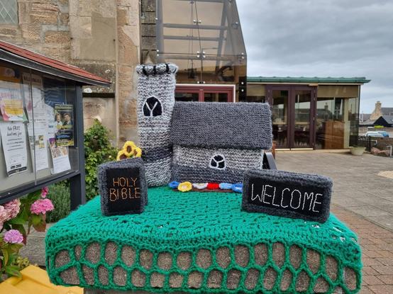 A photo of a post that has been yarn bombed with a knitted church scene. There is green grass on which is a knitted church in shades of grey , a holy bible with a black cover and gold title and a white welcome sign on a black background.