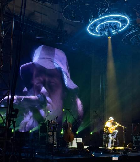 The Billy Strings concert at the Greek Theatre in Berkeley CA on Saturday August 17, 2024. Billy is performing solo, sitting on a stool at the front of the stage playing the Merle Travis song "Muskrat" originally released in 1947.  The video screen behind the stage shows a close-up of Billy's face singing into the microphone with his eyes closed.