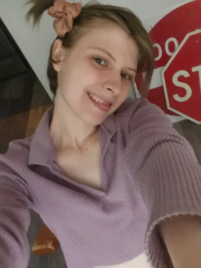 Purple sweater v-neck crop-top with Collar, side-hair pigtail with short blonde-brown hair and Stop & Do Not Enter signs in back corner. White wall and brown and grey floors slanted in picture.