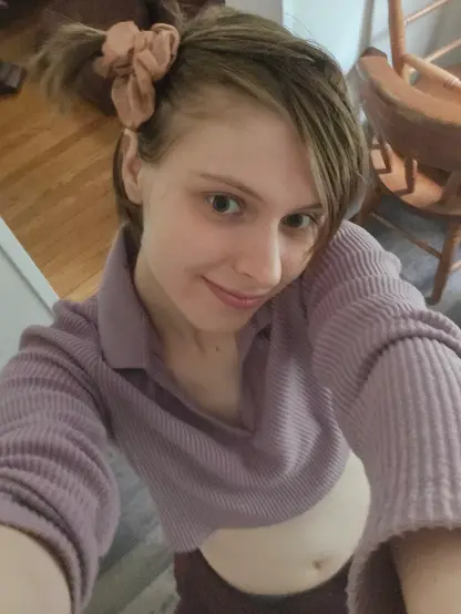 Top-view picture, purple v-neck crop-top, chair and house in background. Blonde Hair tied in side pony with scrunchy. Fuzzy red jacket tied around waist. Arms holding up camera. Dorky side smile, and side-eying the camera.