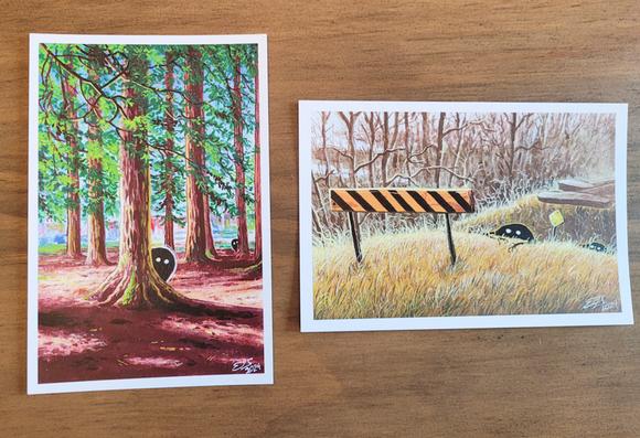 A photograph of two 4 x 6 mini prints. The prints are of paintings depicting small black blob creatures with white eyes. In the first painting, the creatures are in a park peeking out from behind two trees. In the second painting, two creatures are crawling out of the ditch at an abandoned rail crossing.