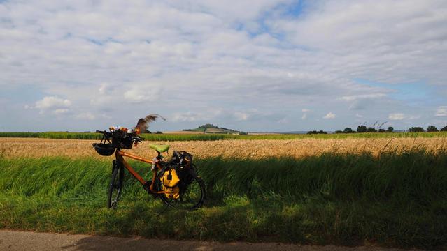Fahrrad bei Sonnenschein am Wegrand vor einem Feld. Blauer Himmel mit weißen Wolken, im Hintergrund ein Berg mit einer Burgruine