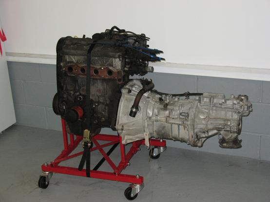 Suzuki G16B 4 cylinder engine sitting on a red engine stand/ dolly with a two wheel drive, five speed manual transmission bolted into the engine and cantilevered above the ground from the engine block.