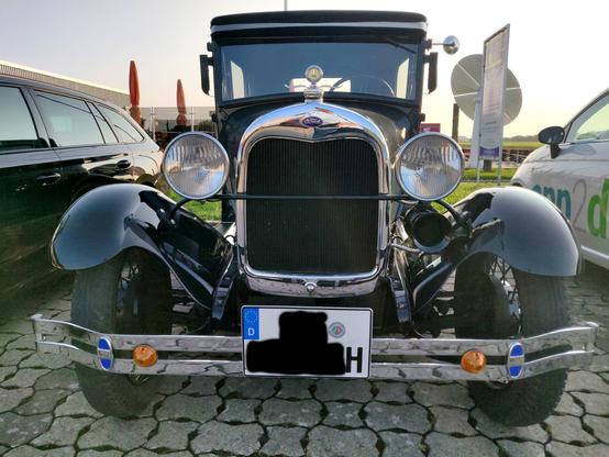 Ein schwarzer Oldtimer, Nahaufnahme von vorn. Ein dunkler Kühlergrill mit Chromrand, zwei freistehende Scheinwerfer, schmale Reifen mit geschwungenen Kotflügeln und ein kastenförmiger Aufbau mit gerader Windschutzscheibe. Ford Model A, Bj. 1929.