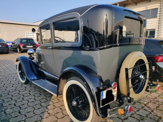 Ein schwarz-grauer Oldtimer von schräg links hinten. Ein kastenförmiger Aufbau mit abgerundeten Ecken, oben schwarz, unten grau. Schmale Weißwandreifen, hinten ein aufgeschnalltes Reservererad. Ford Model A, Bj. 1929.