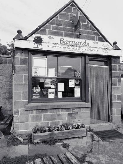 A small stone-fronted shop labeled "Barnards," featuring a sign that states "Purveyors of Locally Sourced Produce." The front has large windows displaying various notices and flyers. There is a planter with flowers at the base.