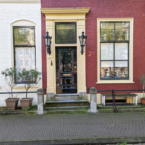 Black door with a large square transom window. There are large lamps flanking the door. The door frame is a light yellow and the door is set on a red wine colored brick wall