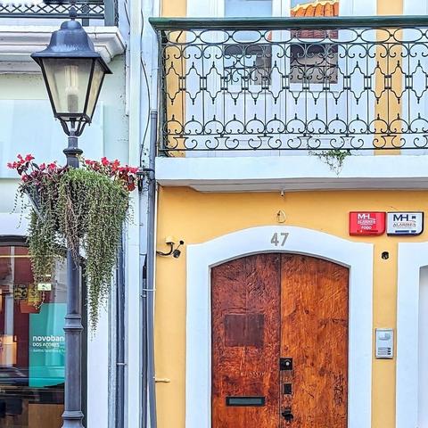 Wooden door with a mottled surface set on a yellow facade. There is a lamppost to the left of the door decorated with a container of pink flowers.
