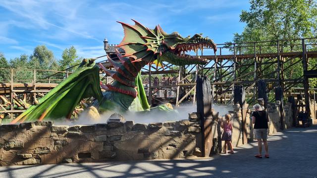 A young girl, accompanied by an older woman, looking up over a clay and brick,  brown wall at an enormous green with red accented statue of a dragon rising out of a mist filled pit in front of a segment of the wooden rollercoaster Joris and the dragon in the Efteling.