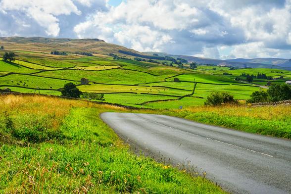 Photo of a green sunny landscape with hills. You see the walls seperating the meadows as dark lines. In the lower right you see a road. The sky is full with white clouds.