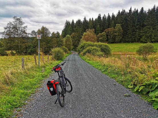 Ein naturgebundener, feingeschotterter Fahr-/Radweg. Auf dem Weg ein Fahrrad mit roter Lenkertasche und Satteltasche. Links und rechts Wiesen und Weiden. Im Hintergrund Bäume und ein grau bewölkter Himmel.