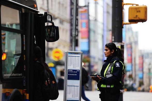 A uniformed female transit police officer watching people boarding a bus in a city environment. The background is blurred but of high-rise buildings
