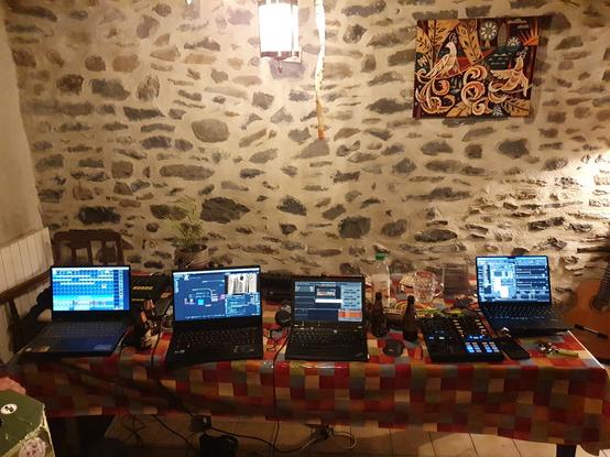 une table avec une nappe old-school devant un mur en pierre auquel est accroché une tapisserie représentant des oiseaux colorés.
Sur la table, 4 ordinateurs et contrôleurs tracktor. un grandma wing a peine visible