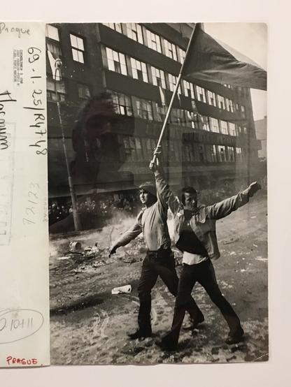 black-and-white photo, vertical orientation:

two young men walk along a desolate streetscape in front of a modern building, one defiantly holding aloft the national flag

various press stamps along left margin