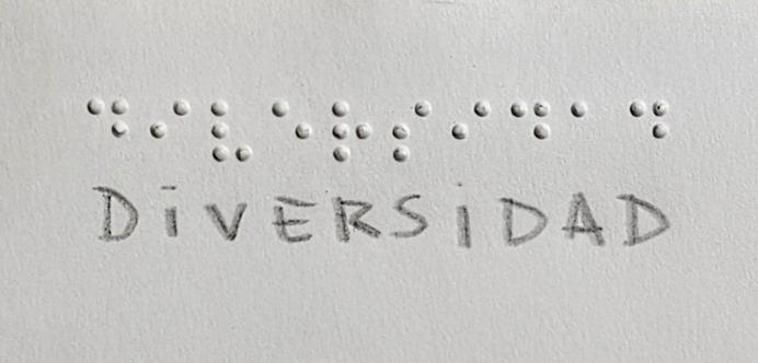 La imagen muestra la palabra "DIVERSIDAD" escrita en dos formas: en la parte superior está en braille, y justo debajo está escrita en letras mayúsculas a mano. El fondo es de color claro, probablemente blanco o gris claro.