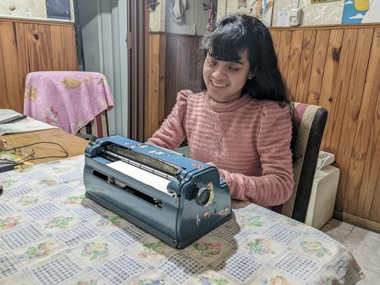 La imagen muestra a una niña sentada en una mesa, sonriendo mientras utiliza una máquina Perkins Brailler, que es una máquina de escribir en braille. La máquina es de color azul y tiene algunos adhesivos decorativos. La niña lleva una camiseta de manga larga de color rosa y tiene el cabello largo y oscuro. La mesa está cubierta con un mantel con un diseño de cuadros y flores. Al fondo, se puede ver una pared de madera y una puerta entreabierta. También hay una silla cubierta con una manta rosa con dibujos.