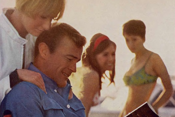 1968 

The image depicts a sunny outdoor scene where several individuals are interacting with each other on what appears to be the deck of a boat or near water.
There is one person seated and smiling, being assisted by another who is standing beside them, seemingly helping them stand up or move.
In the background, there are three more people observing this interaction; one woman is looking towards the camera while two others appear to be facing away from it.
The setting suggests a casual, leisurely atmosphere with clear skies and bright sunlight.
Tags: boat, water, people, assisting, outdoors, smiling, standing