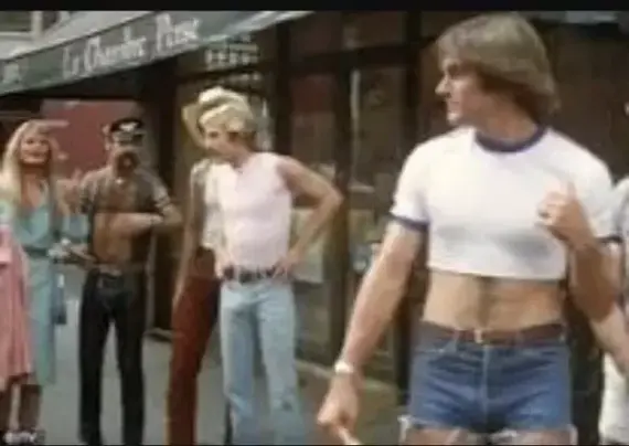 A group of people are standing on a street in what appears to be a scene from the 1970s. One man in the foreground is wearing a tight white crop top and very short denim shorts, drawing attention from the others around him. The group includes individuals dressed in various styles typical of the era, including a man in a leather jacket and another in flared jeans. The overall vibe is retro, with a focus on fashion trends from that period.