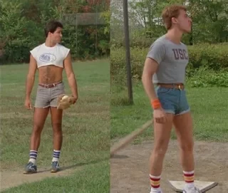 Two men are standing on a grassy field, dressed in tight, vintage athletic wear. The man on the left is wearing a cropped t-shirt, short denim shorts, striped socks, and holding a baseball glove. The man on the right is also wearing short shorts, a fitted t-shirt with "USC" printed on it, and striped socks, and is holding a baseball bat. Both men are styled in a way reminiscent of 1980s athletic fashion.