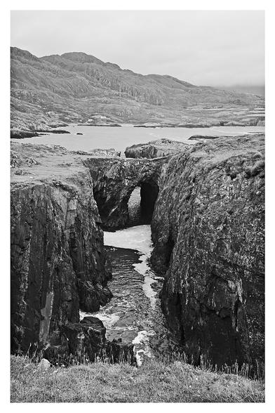Sea arch along the coast near Allihies. Looking back over the inlet towards Knockgour hidden beneath a blanket of cloud. 

On the ridge line, where it slopes down, to the upper right, the remnants of the engine house of the mountain mine is just visible below the clouds.