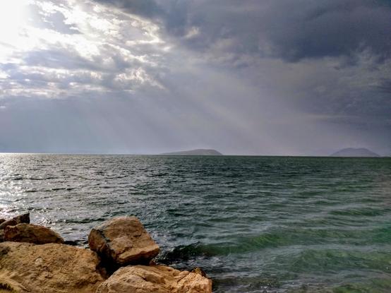 Der Blick geht über den Golf von Ambrakia zu zwei kleinen Inseln, die verschwommen am Horizont aus dem Wasser ragen. Im Vordergrund links befinden sich kleine Felsbrocken des typischen rotbraunen Steins dieser Gegend. Das Meer ist leicht gewellt und wird von der Sonne, die am bewölkten Himmel links oben durch die Wolken bricht, beschienen.

The view extends across the Gulf of Ambrakia to two small islands that rise hazily out of the water on the horizon. In the foreground on the left are small boulders of the typical reddish-brown stone of this area. The sea is slightly undulating and is illuminated by the sun, which breaks through the clouds in the overcast sky at the top left.