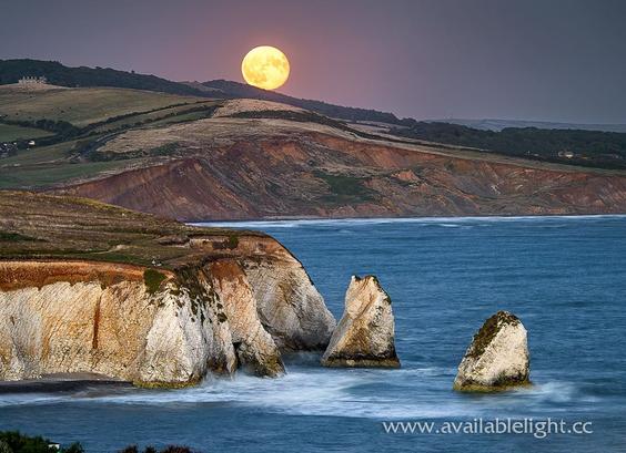 An almost full moon emerges from behind a cliff over the sea. The coastline has several large rock formations in the deep blue sea. 
I'ts almost nightfall, the moon quite yellow in appearance as it moves through the atmospheric haze.