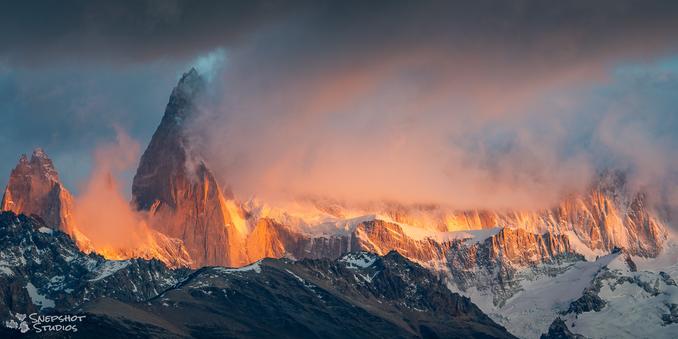 Wide-screen photo of a rugged mountain range. Some glaciers are visible. The mountain tops are hidden in low cloud. The side of the mountain below the cloud layer is hit by strong orange sunrise light.