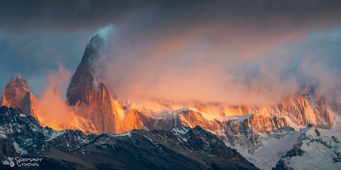 Wide-screen photo of a rugged mountain range. Some glaciers are visible. The mountain tops are hidden in low cloud. The side of the mountain below the cloud layer is hit by strong orange sunrise light.