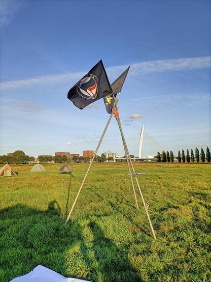 Picture of the camp with two flags, the antifa flag and the refugees welcome flag. Behind the flags you see a few tents and behind that the built-up area of Utrecht and the Prins Claus Bridge across the Amsterdam–Rhine Canal.