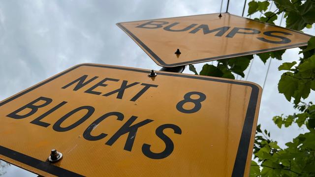 a yellow caution street sign, shown at a close up at a steep angle, reads BUMPS next 8 blocks