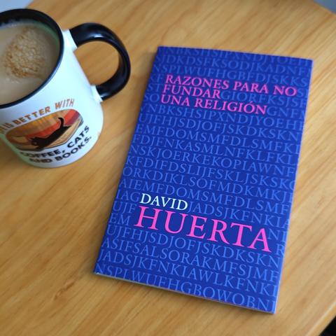 Sobre una mesa de madera, una taza llena de café y un ejemplar del libro "Razones para no fundar una religión" de David Huerta.