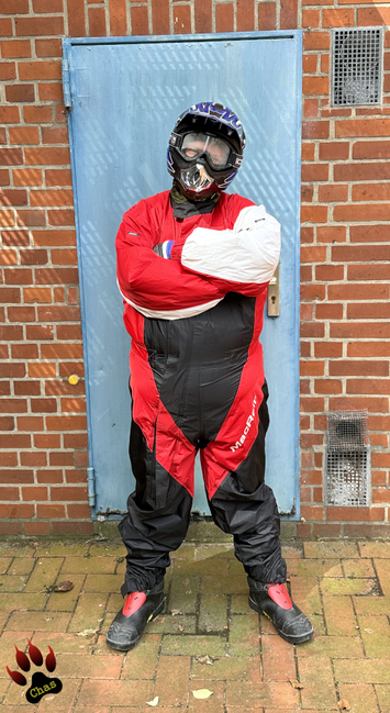 person wearing a red/black/white motorbike rainsuit, Haix boots and a motocross helmet standing outdoors in front of a brick wall with a blue door, arms crossed