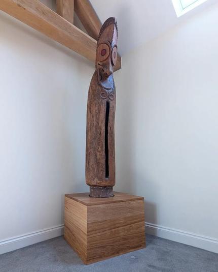 A wooden sculpture (actually a musical instrument and communication device) made from a tree trunk stands in the corner of a room. It's been carved to look like a long, stylized face: large eyes, a prominent nose, a sort of moustache, and a long slit which is actually the mouth, open in song. The eyes have red pupils and there are some possible facial tattoos in black, not otherwise the wood is unpainted. It stands in a beautifully crafted oak plinth. In the periphery are magnolia walls, wooden beams, a bit of a window, and a grey carpet.