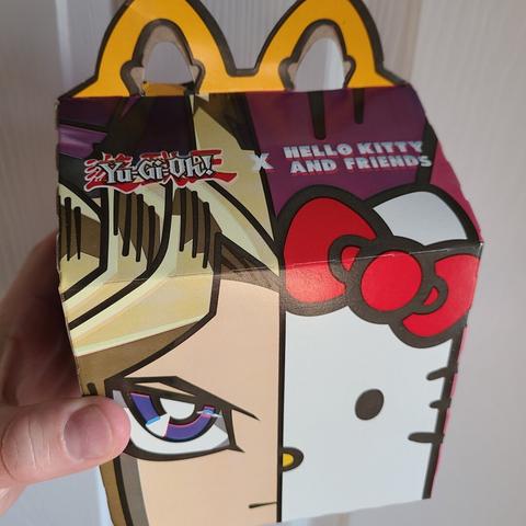 Photograph of a McDonald's Happy Meal box. The box has the traditional yellow arch on top, but the side of the box shows a split with half of Yugi from Yu Gi Oh's face (left) next to Hello Kitty's face (right). Above Yugi's face is the Yu Gi Oh game / anime's logo. Above Hello Kitty's face is the Hello Kitty logo. There is a relatively centralized X symbol between them.