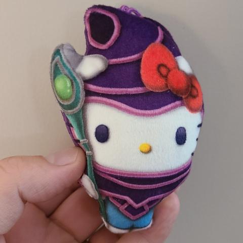Photograph of an adult white person's hand holding a small plush figure of Hello Kitty (white cat with a red bow) dressed as the Dark Magician from Yu Gi Oh (purple suited mage costume holding a large green staff).