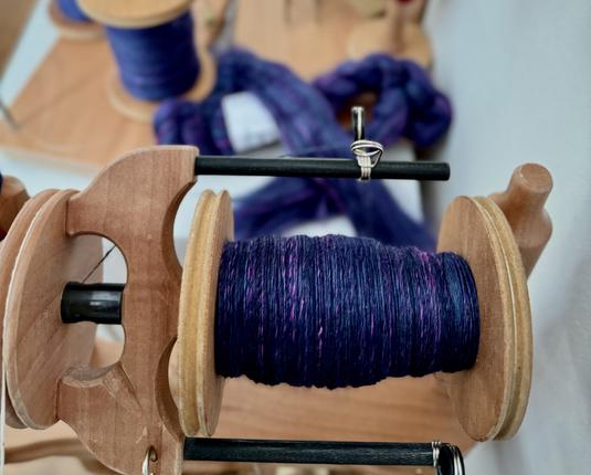 A large wooden bobbin on a spinning wheel fed via a sliding flyer is half-full with a gorgeous dark blue/purple yarn flecked with teal and pink. The forlorn braid and fiber waiting to be spun is sitting behind, near the two full bobbins, and all are wondering why the spinner has seemly forgotten them.