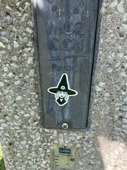 On a metal rectangle on a concrete post, a sticker of a young witch with a black hat and an intense look- drawn in black and white.