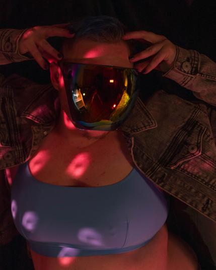 A white person with blue and brown hair and dark body and facial hair in a colorful reflective face shield, a moto jean jacket, a blue bra, and short shorts posing in disco lights and colored lights.