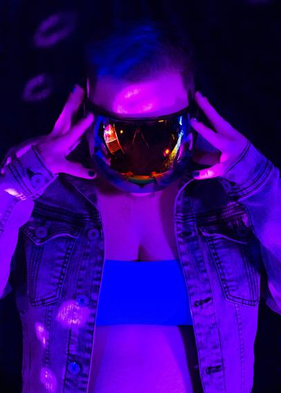 A white person with blue and brown hair and dark body and facial hair in a colorful reflective face shield, a moto jean jacket, a blue bra, and short shorts posing in disco lights and colored lights.
