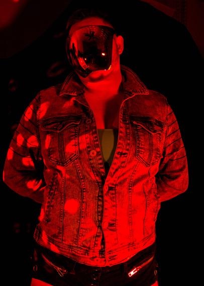 A white person with blue and brown hair and dark body and facial hair in a colorful reflective face shield, a moto jean jacket, a blue bra, and short shorts posing in disco lights and colored lights.