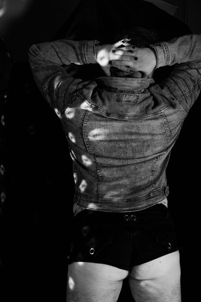 A black and white photo from behind of a white person with blue and brown hair and dark body and facial hair in a colorful reflective face shield, a moto jean jacket, a blue bra, and short shorts posing in disco lights and colored lights.