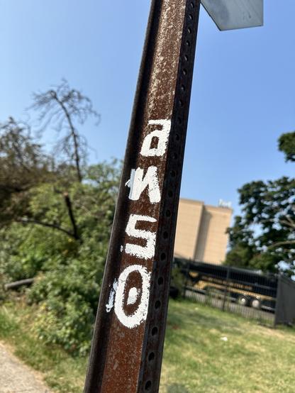 ANSO in vertical white block letters on a rusty streetsign post, with grass and bushes and a tan building and blue sky in the background